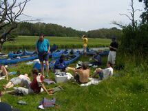 People having lunch in the grass with canoes in the background. Photo. 